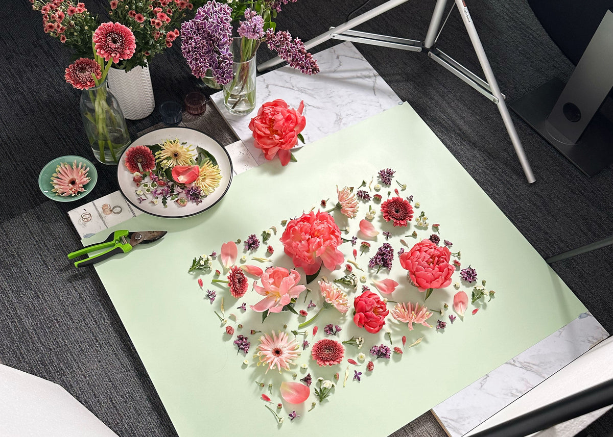In the studio behind the scenes peek at a flower flatlay photo shoot by floral art photographer Connie Chan. Flower on set are coral peonies, gerberas, lilacs and other mixed blooms on a pale green surface.